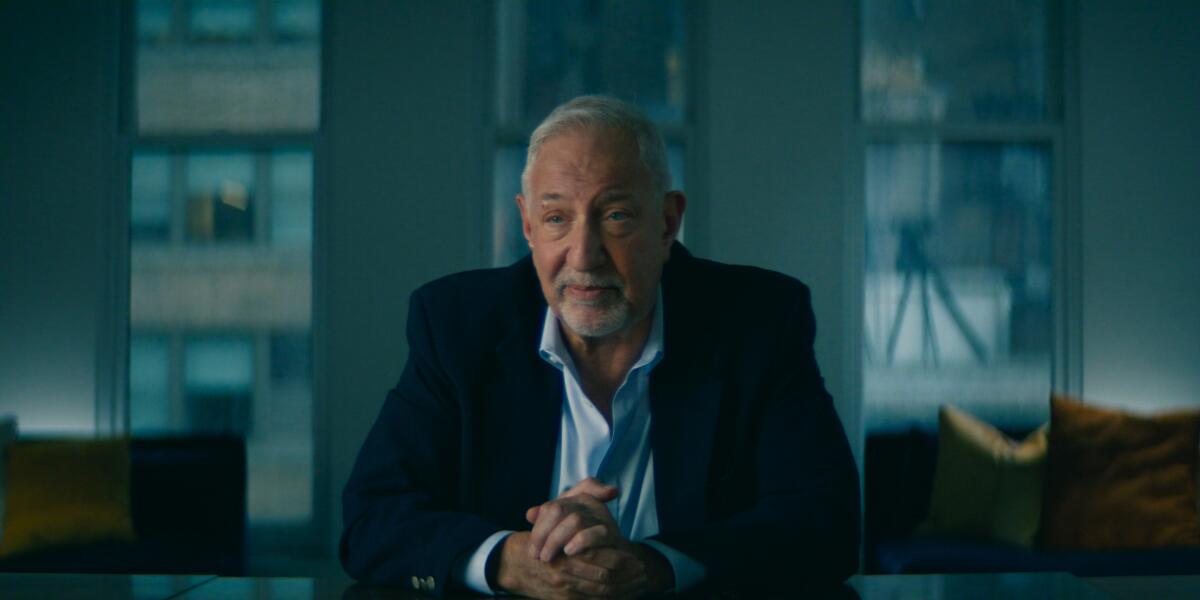 A man with a dark blazer sits with his hands clasped.