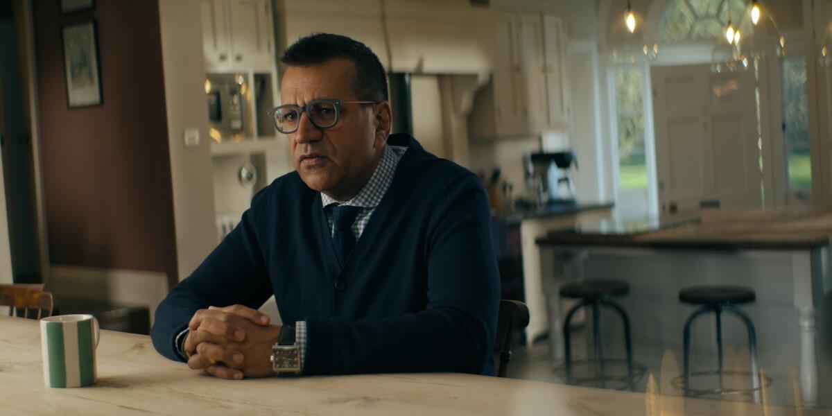 A man in glasses sits at a table counter with a coffee cup near him.