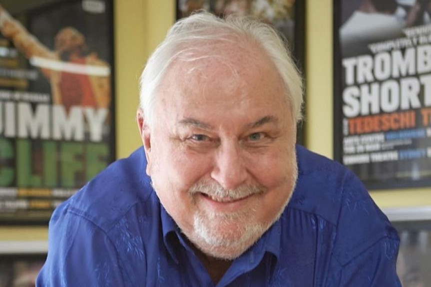 A grey haired man leans forward towards the camera with a broad grin.