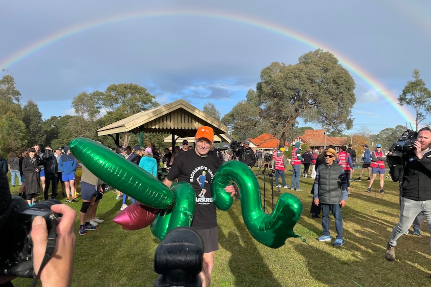 A man wearing an orange cap and black t-shirt stands in a park while holding green balloons with a rainbow in the background.