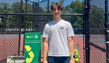 A young man stands beside a green recycling bin for used tennis balls at a tennis court.