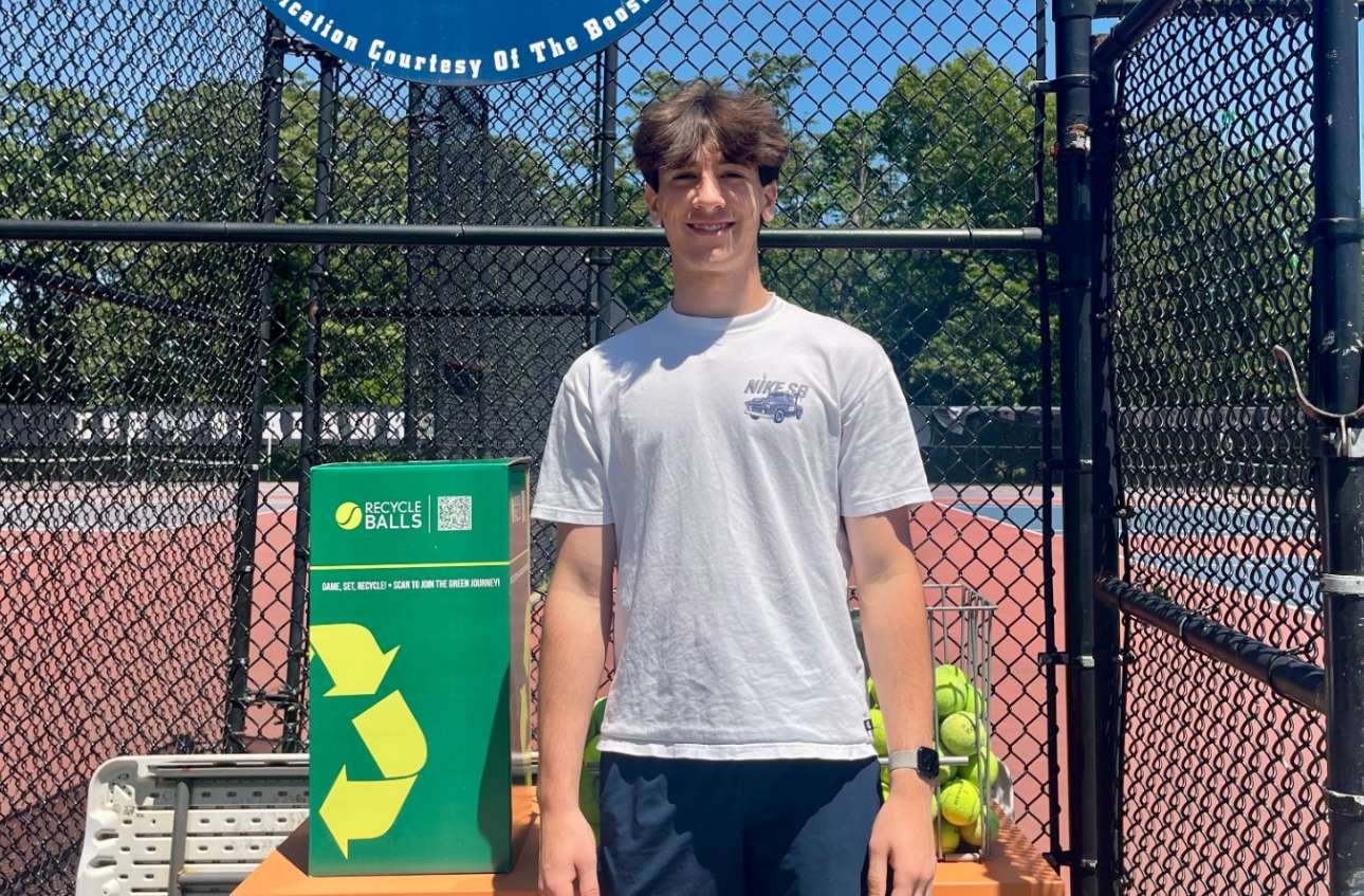 A young man stands beside a green recycling bin for used tennis balls at a tennis court.
