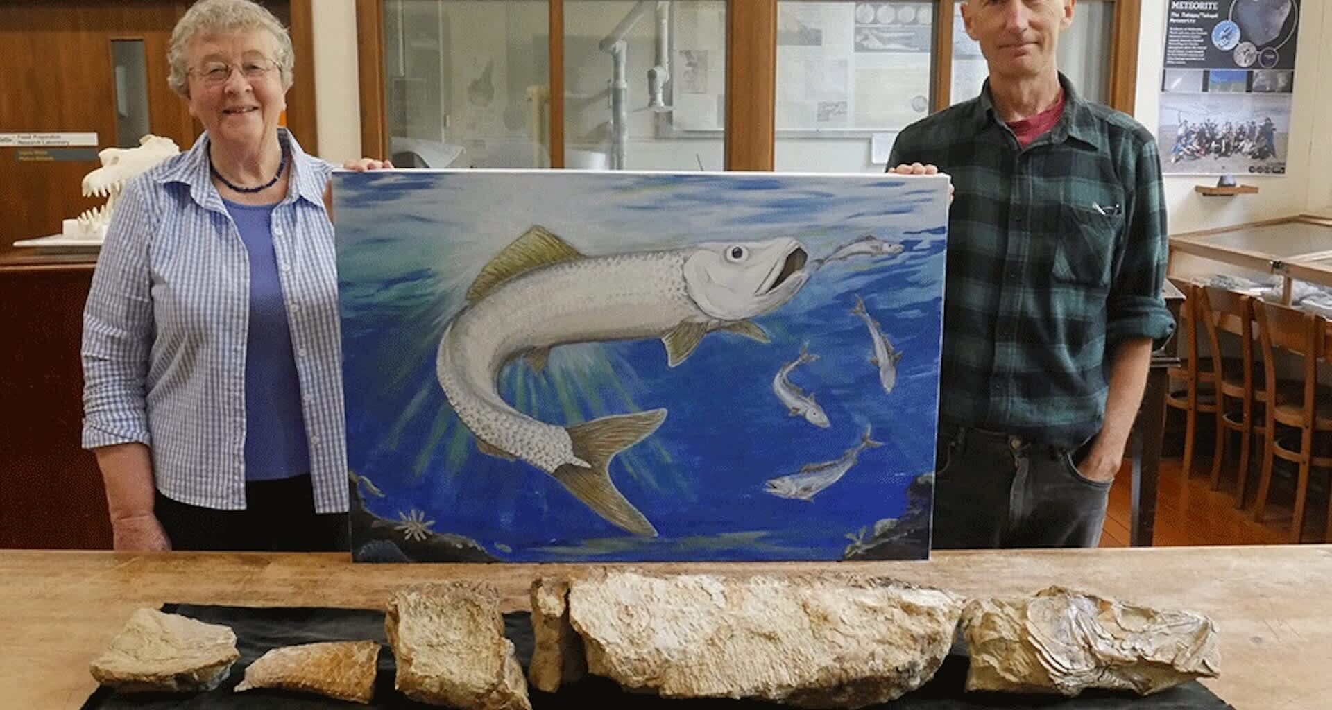 A woman and a man stand beside a painting of a fish, with fossilized fish remains displayed on the table in front of them.