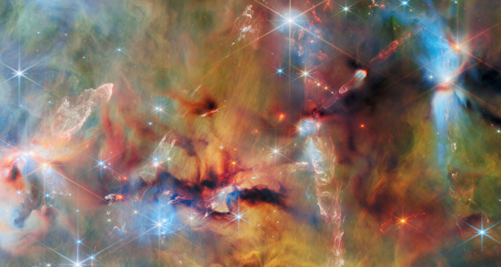 An area inside a star-forming molecular cloud. The background is covered with layers of gas and dust in blue, green and yellowish colors. Thicker clumps of cold dust, dark brown to black, block out light completely. Stars lie among and atop the clouds, from small orange ones to large white or blue ones. Waves and streams of glowing whitish gas are created by jets from protostars colliding with the surrounding material.