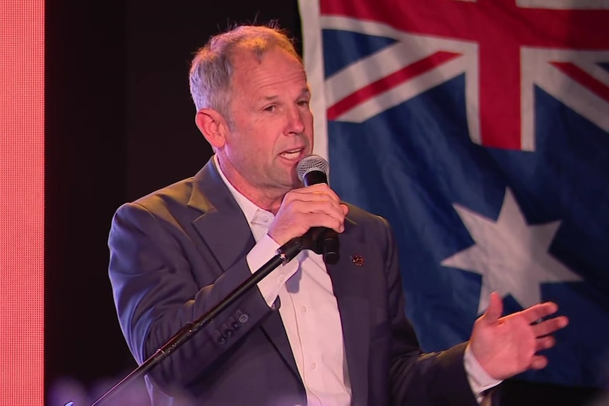 Rod Caddies speaking with a mic in front of an Australian flag.