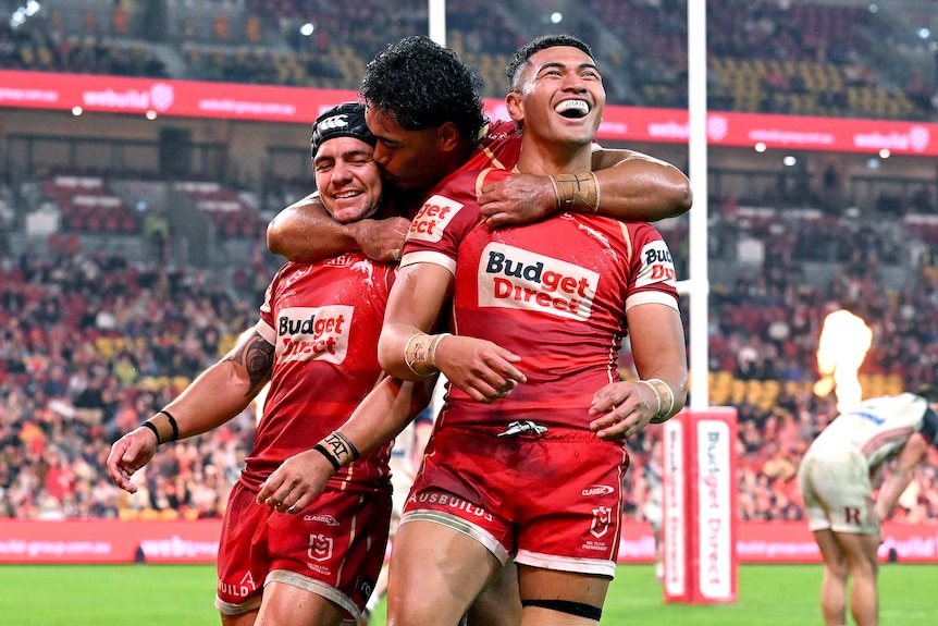 A rugby league player in red is hugged by his teammates while celebrating a try