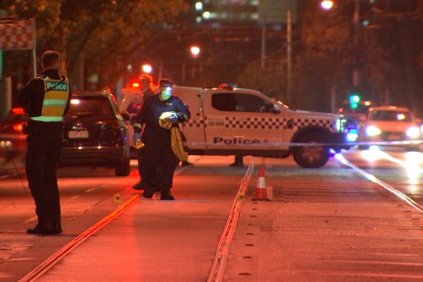 A police officers standing in the middle of a road with a detective walking in front of a police car and police tape.