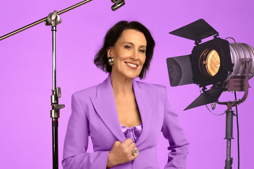 Virginia Trioli, wearing a fitted blazer, poses for a photo in a studio next to a spotlight and microphone