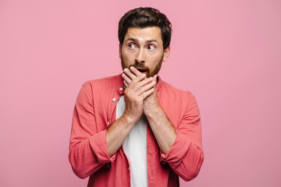 Young bearded man covering his mouth with hands, expressing fright, looking to side