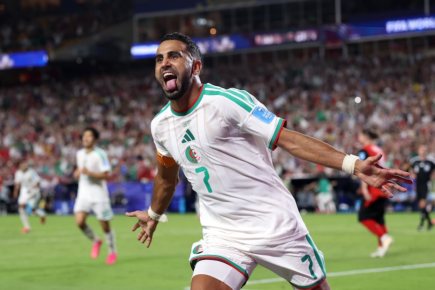 A soccer player in white runs with arms outstretched and tongue poked out in celebration