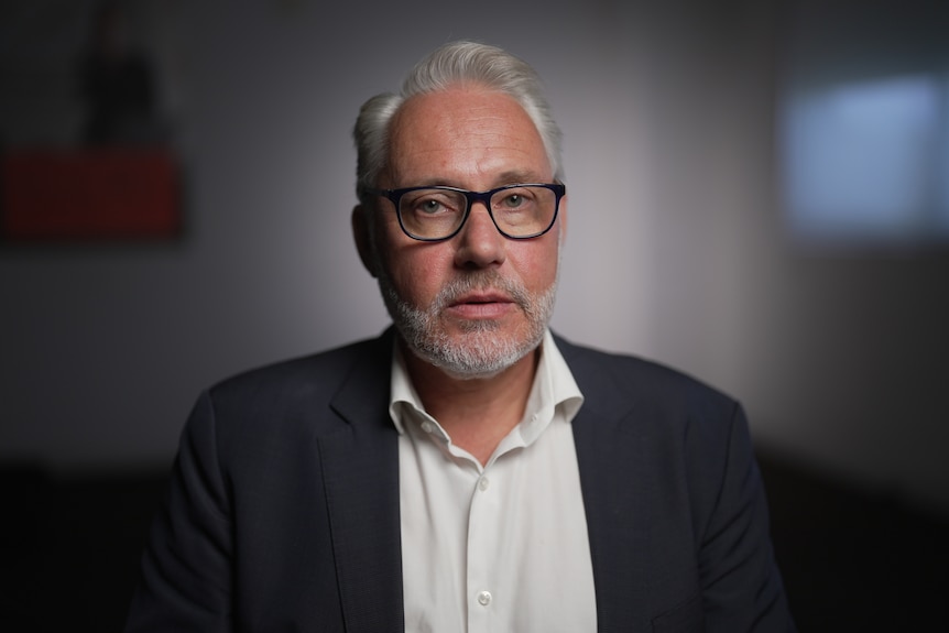 A man wearing a suit jacket and glasses looks at the camera he is inside a room. His expression is neutral.