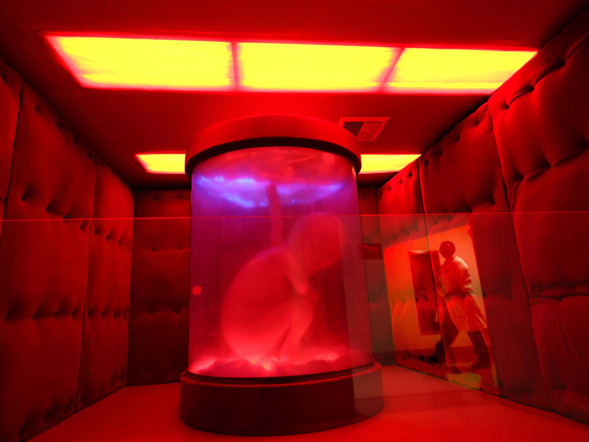 A person in a white coat passes a room with a red-lit fetus in a jar.