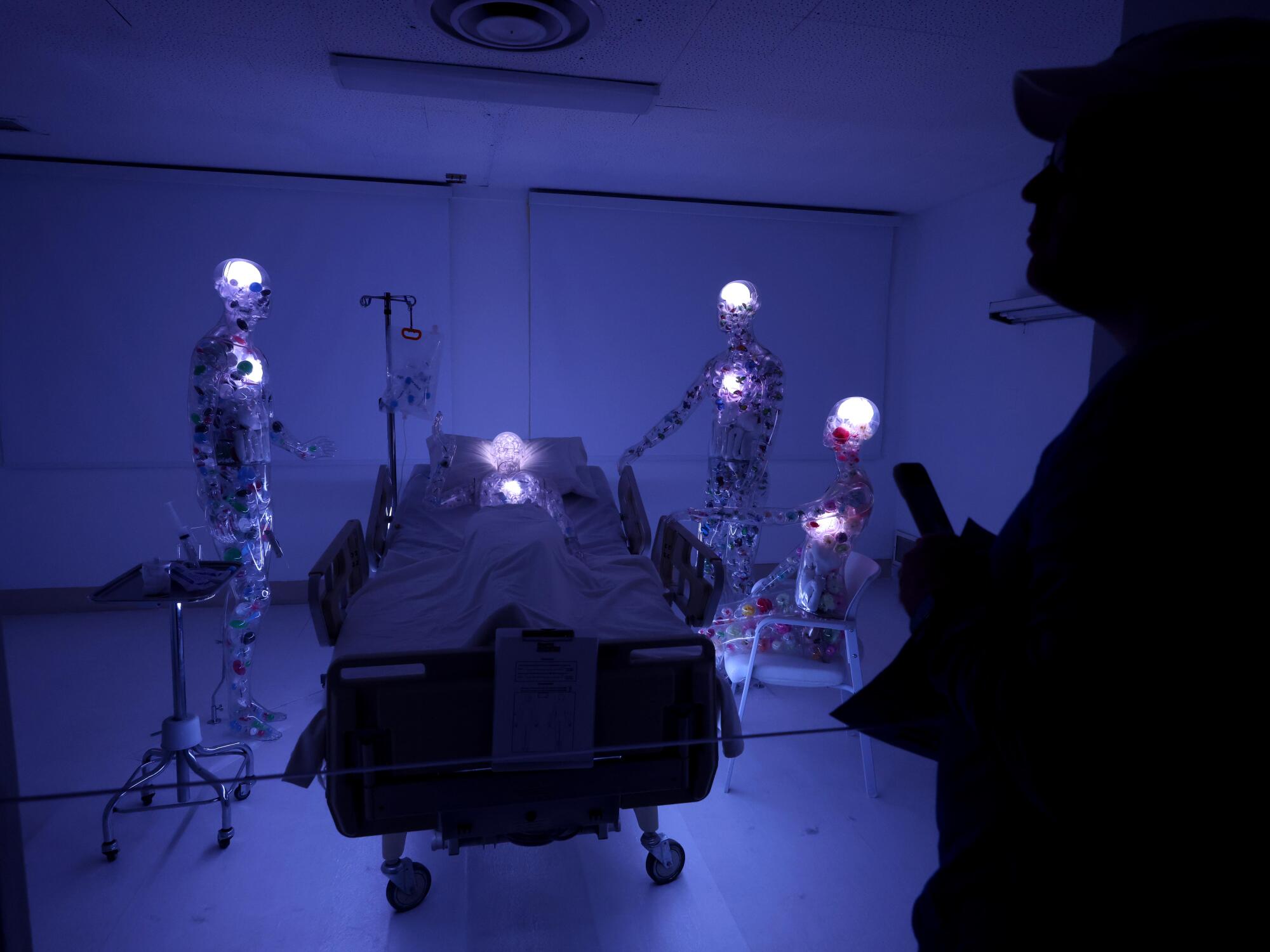 Illuminated figures stand around a hospital bed.
