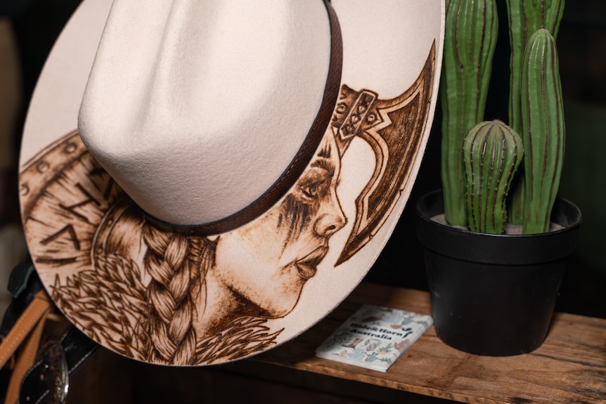 Cowboy hat with women's face and axe draw on its rim next to a potted cactus plant