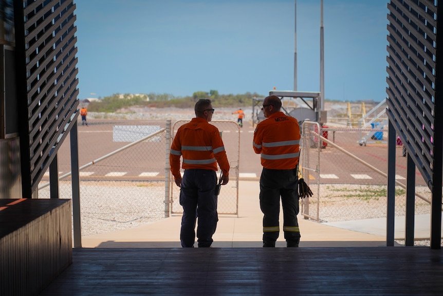 workers in hi vis stand in the shade of a shed overlooking the airstrip