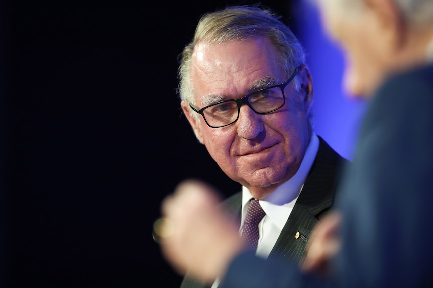David Gonski with grey hair and black-rimmed glasses, and smiling slightly, turns his head to face a person just visible nearby