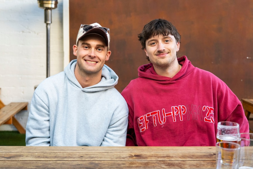 A man in a cap and grey hoodie smiles sitting next to his friend in a red jumper with a cup of beer in front of him.
