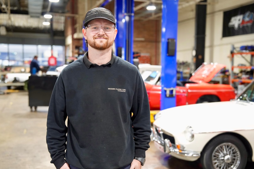Nick in a hat and glasses in front of vintage cars