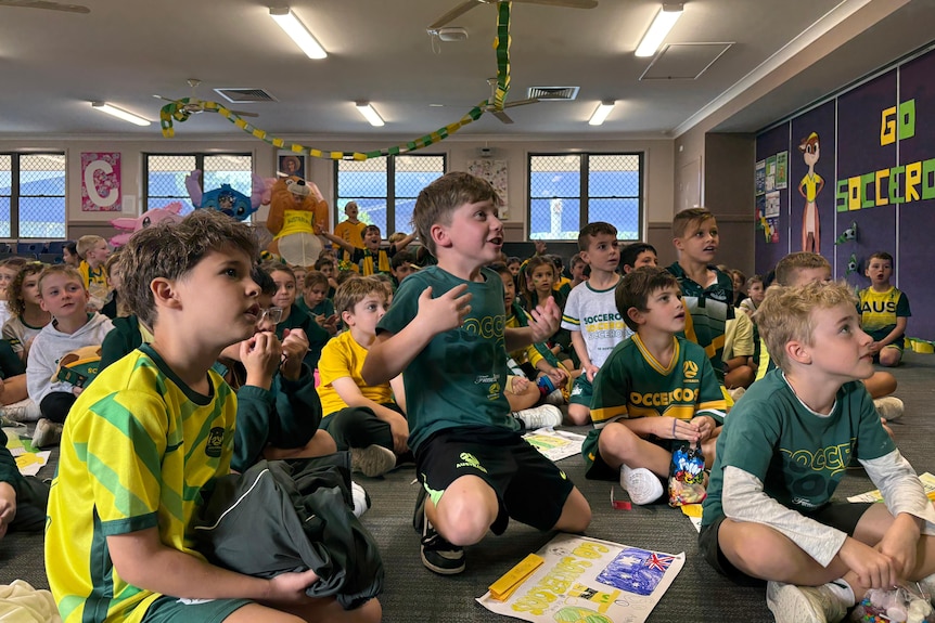 School kids dressed in green and gold sit on the floor watching the game in a classroom.