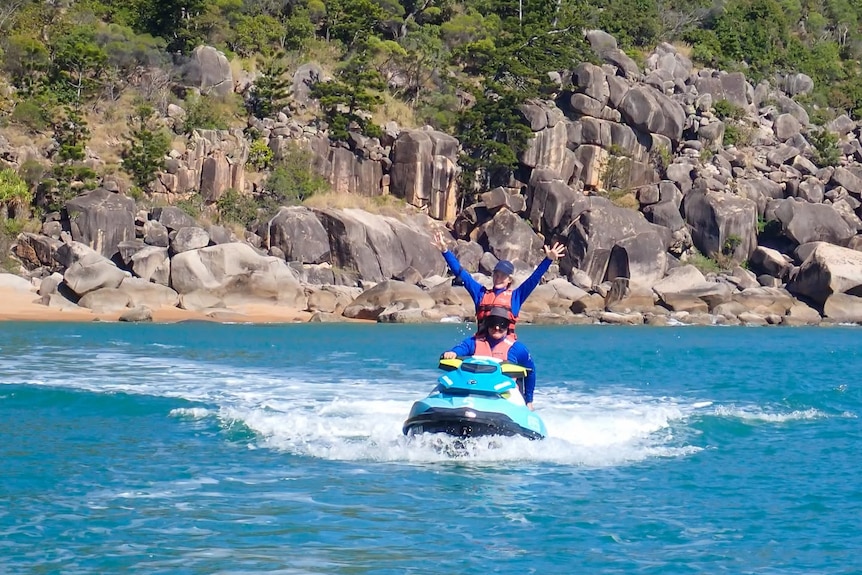 A man sits on a jet ski driving it, with a woman standing on the same jetski behind him with her hands in the air