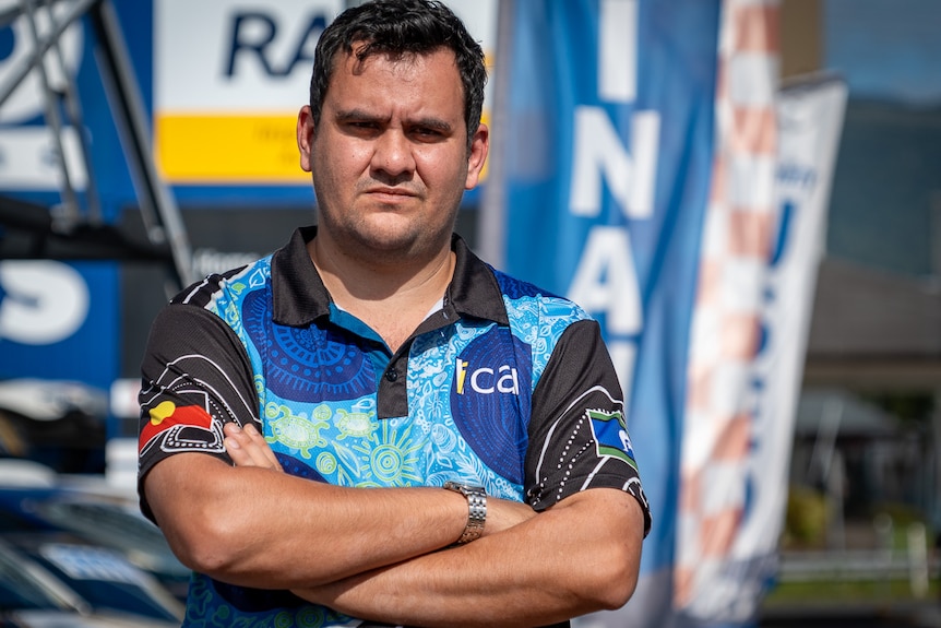 A man in a polo stands at a used car lot with colourful flags in the background.