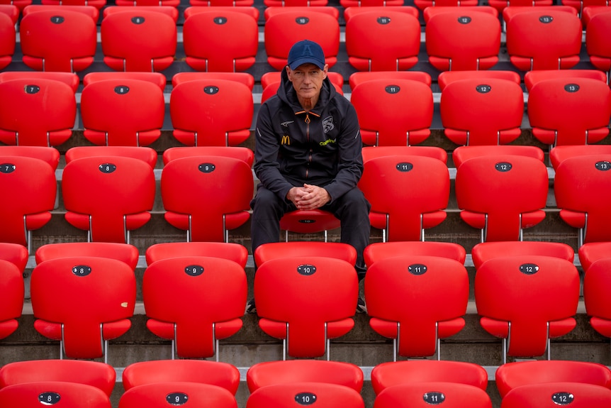 A man in a cap sits in the audience seats overlooking a soccer field with AUFC emblazoned in the audience stand