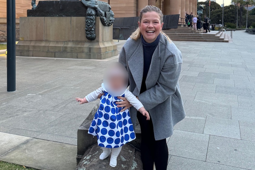 leah stewart smiles at the camera while holding the hands of her baby daughter outdoors
