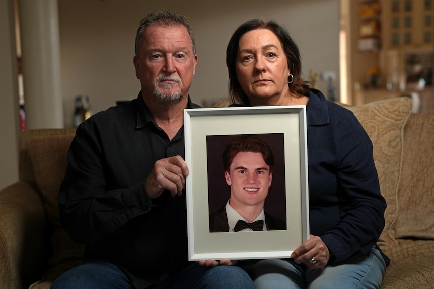 A man and woman sit on a couch in a living room holding a photo of a smiling young man. Their expressions are serious.