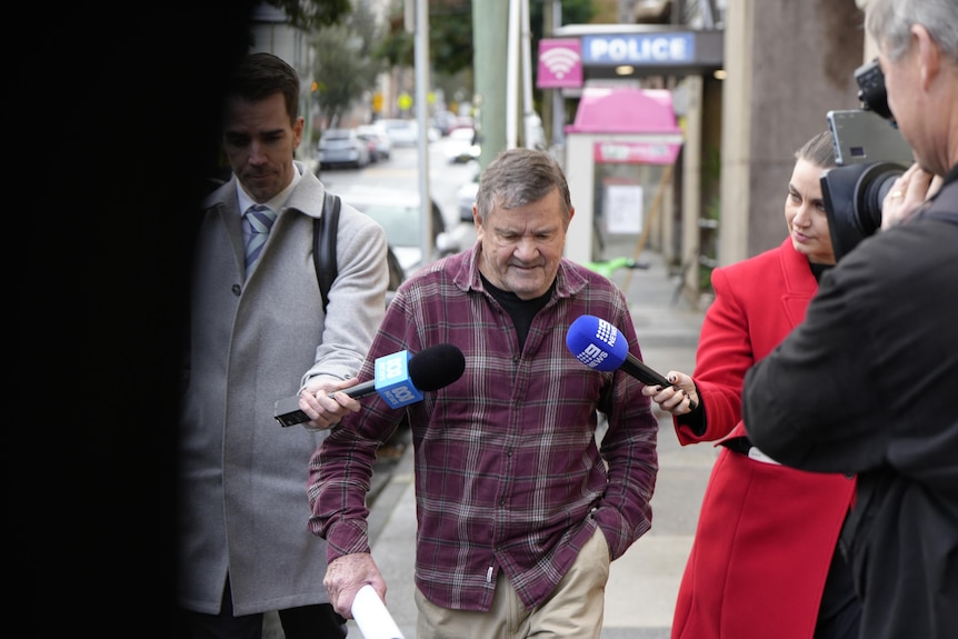 Mr Cribbin in a black undershirt and chequered red longsleeve is followed by reporters as he walks.