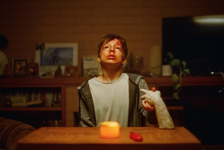 A white young man with brown hair, a bloodied face and dilated pupils, is sitting petrified at a kitchen table.