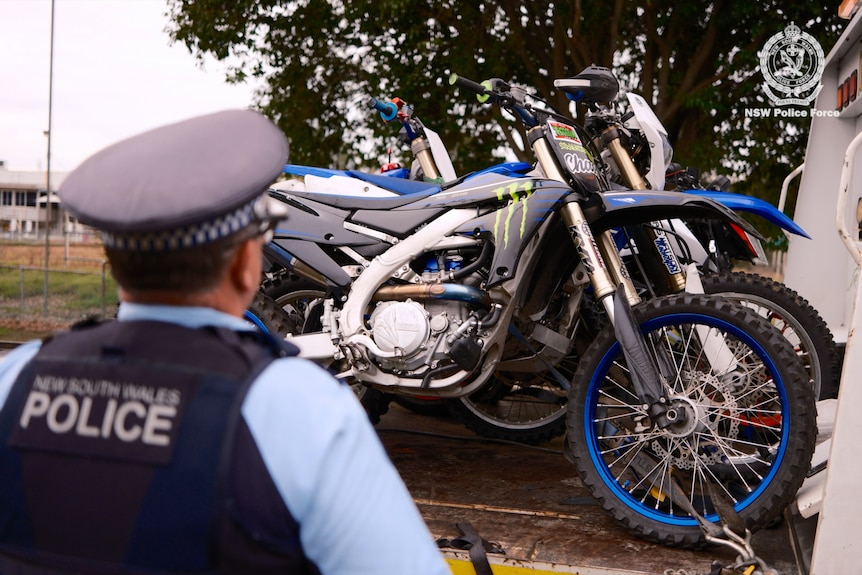 A police officer standing in front of two motorbikes on the back of a tow truck.