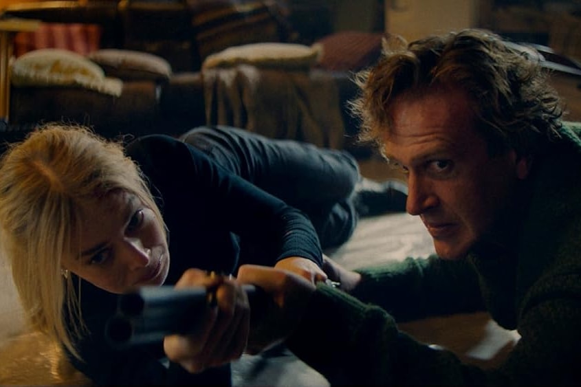 A man and a woman lie on the floor of a house fighting over a gun.