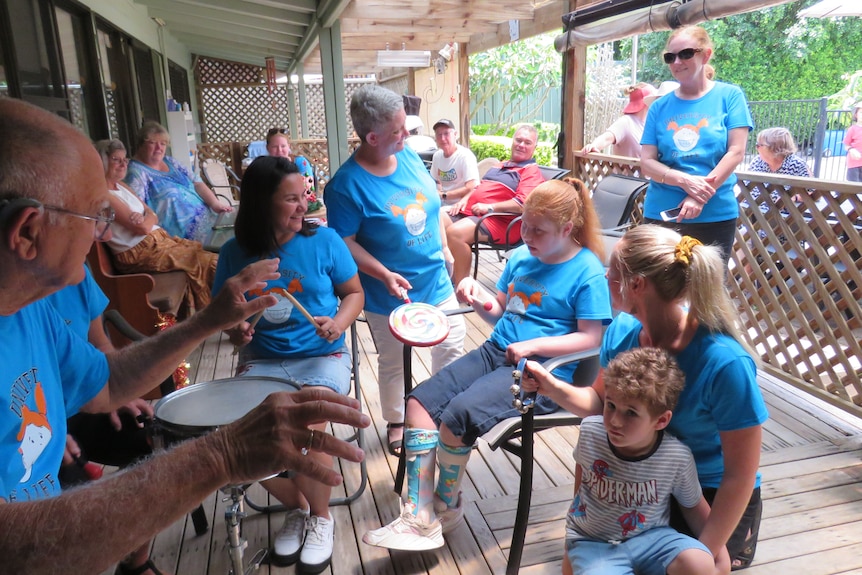 Many people sitting around Lillian wearing blue UOL T-shirts - Lillian, - red hair, holds a small frame drum and stick beater