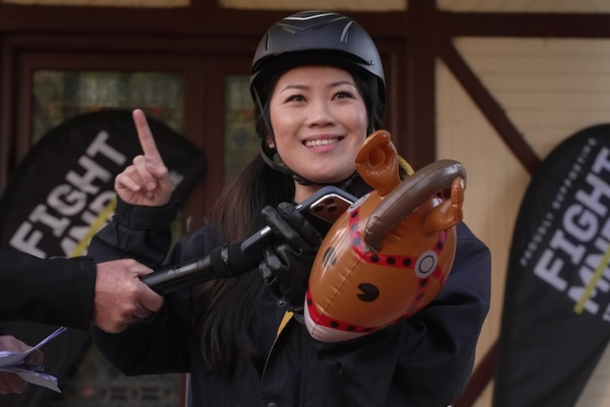 A woman wearing a black helmet holds a hobby horse in one hand and her finger up in air.