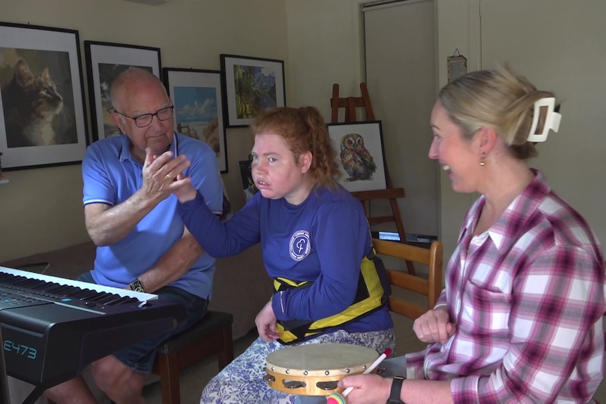 A young woman with a disability high fiving her pop at the piano keyboard with her support worker holding a tambourine