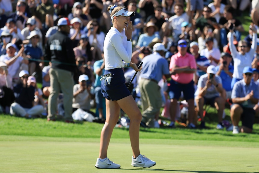Nelly Korda laughs, holds a hand to her mouth, holds club in other hand as people watch on, blurred, green grass, wears white t.