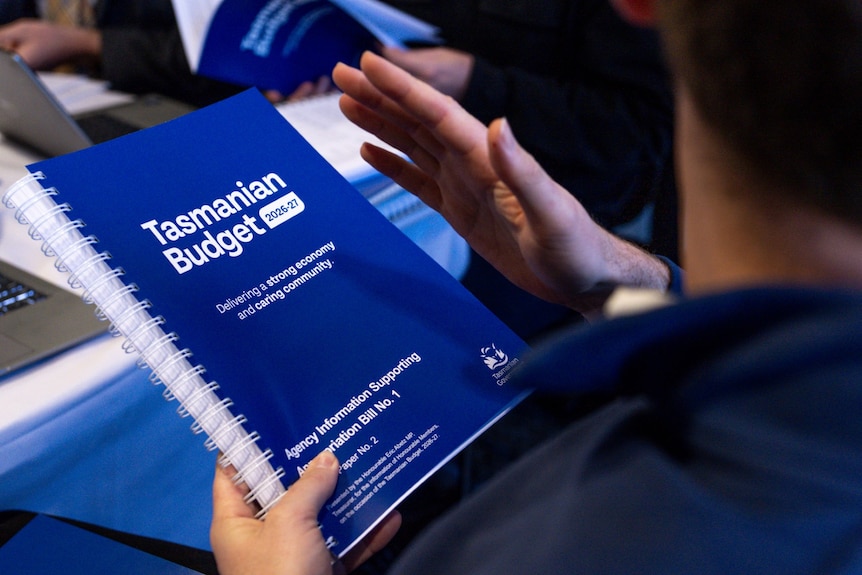A photo taken over the shoulder of a male holding the blue Tasmanian Budget book.