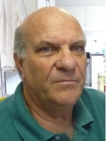 A headshot of a older man wearing a green T-shirt, in what looks like a laboratory.