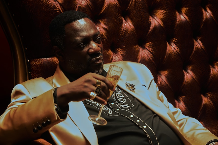 Actor playing Alamo sitting down with a champagne flute and stern expression on face, gold suit jacket, black shirt