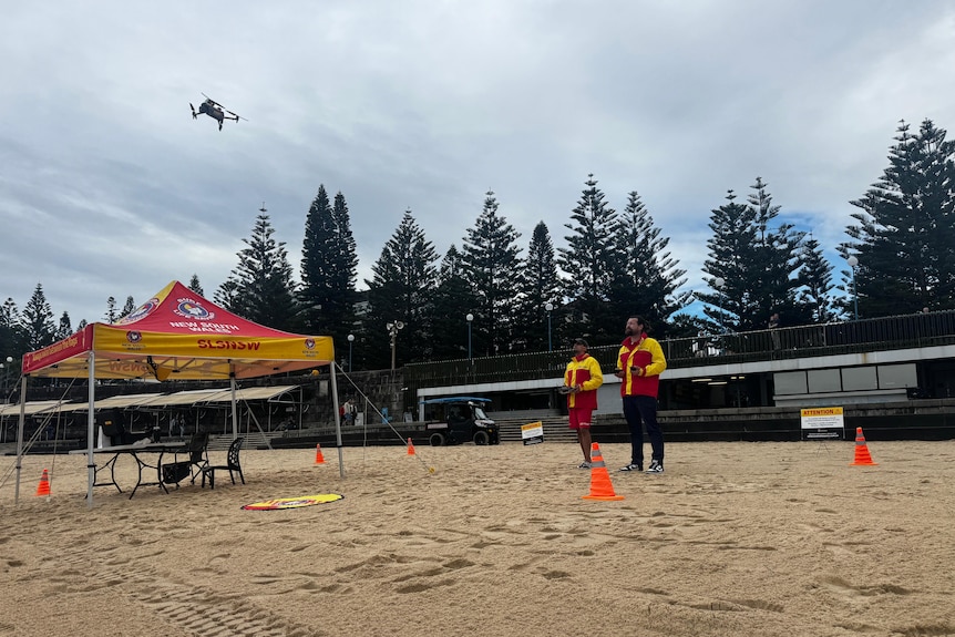 Two surf lifesavers monitor and control a drone on a beach near a tent with a table and chairs beneath it.