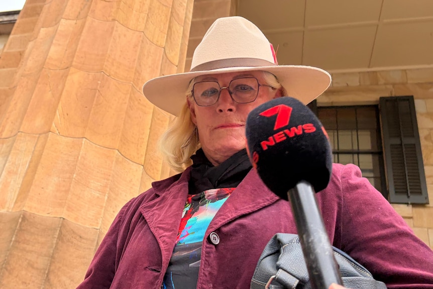 A person wearing an Akubra hat and maroon jacket with a 7 news microphone near their face in front of a stone building