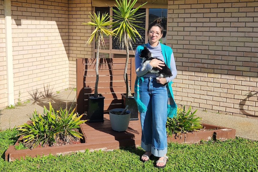 A woman holding a dog, standing in front of a garden at the front of her home.