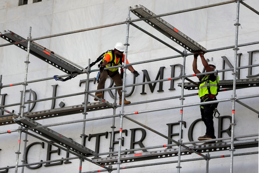 Two men in yellow hi-vis clothing and white hard hats stand on scaffolding in front of a large metal TRUMP sign.