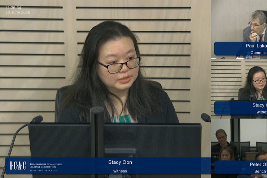 A dark-haired woman in glasses sits behind a computer screen and some microphones.