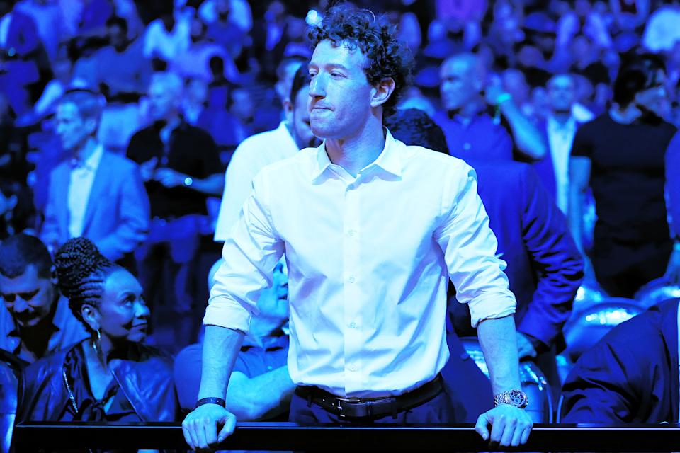 A man with curly hair wearing a white button-up shirt with rolled-up sleeves stands with his hands resting on a railing. He is looking to his left with a thoughtful expression. The background is filled with a crowd of people, all bathed in blue lighting.
