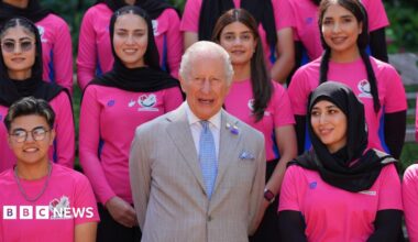 King Charles wearing a pale grey suit with white shirt and blue tie, with members of the Afghan women's cricket team - wearing hot pink T-shirts - on either side and behind him. All are looking ahead and smiling