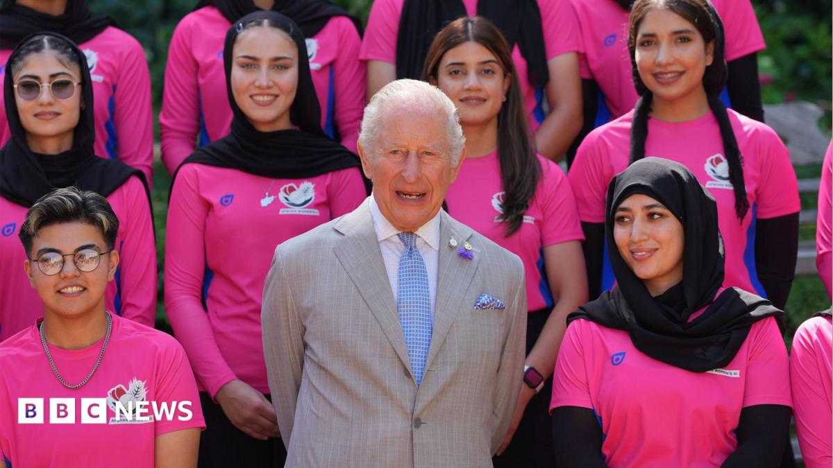 King Charles wearing a pale grey suit with white shirt and blue tie, with members of the Afghan women's cricket team - wearing hot pink T-shirts - on either side and behind him. All are looking ahead and smiling