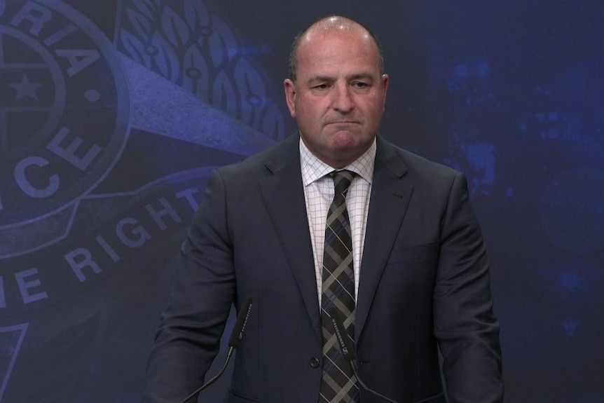 A middle-aged man in a dark suit stands in front of a Victoria Police backdrop.
