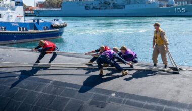 Five men in military uniforms and brightly colored vests attach mooring lines to a submarine in a bay.