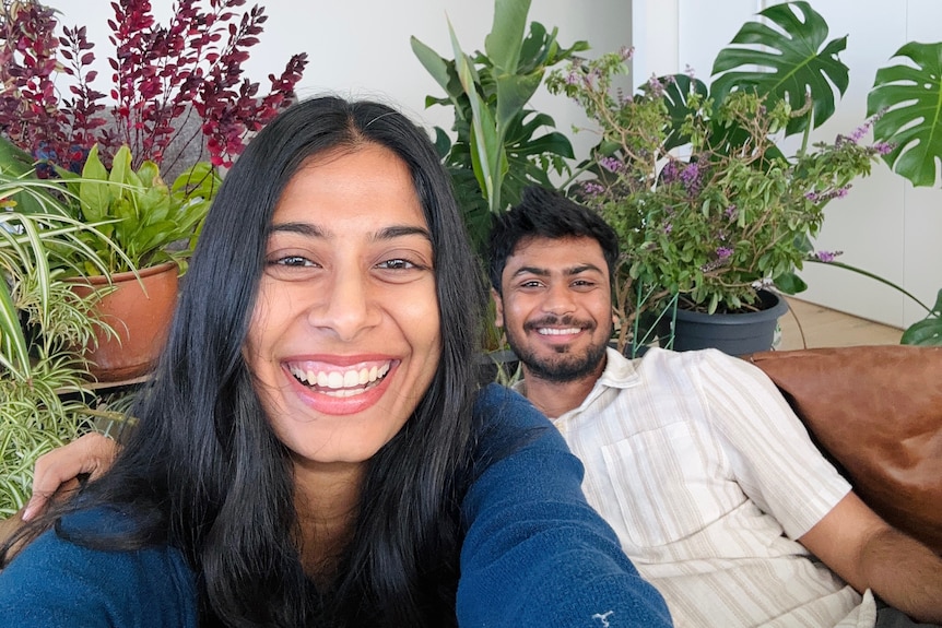 A young woman with long hair takes a selfie with a young man in the background